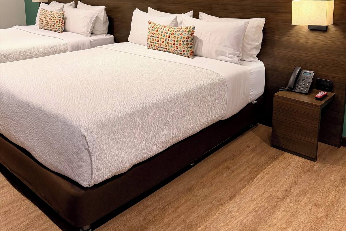 A hotel room with two beds, a large abstract painting on the wall, a wooden headboard, and a bedside table with a phone and a red object.