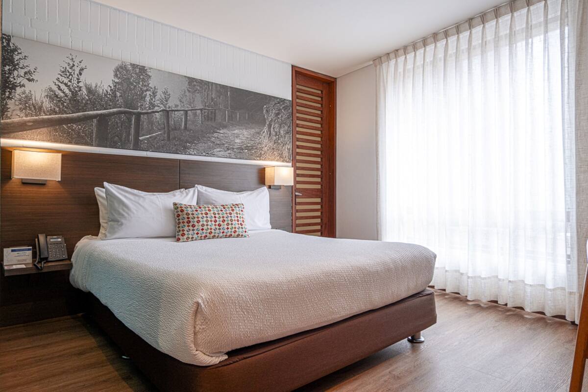 A neatly made bed with a headboard, a bedside table with a lamp, and a large painting on the wall.