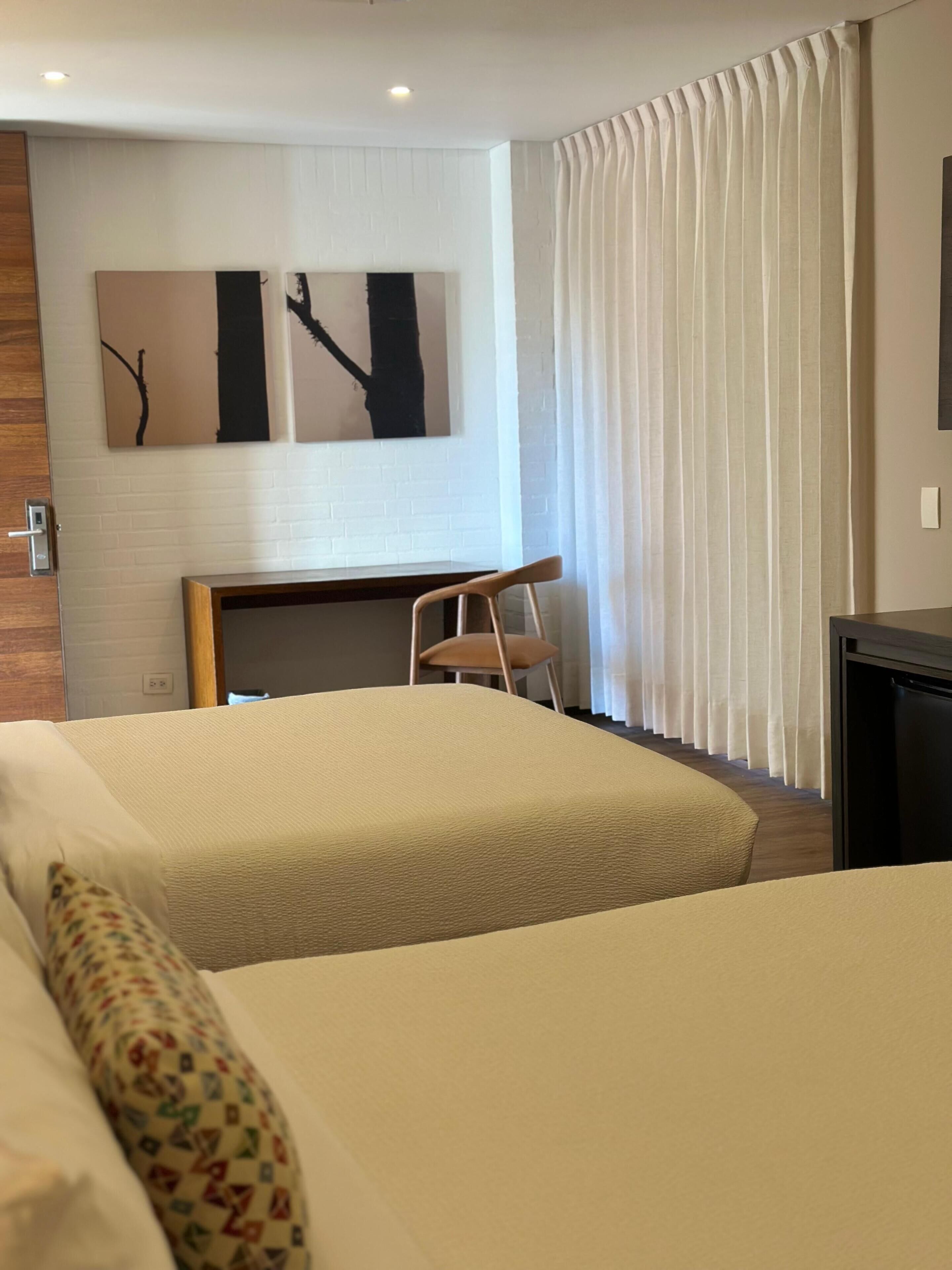 Habitació d'hotel amb dos llits, una taula de treball, una cadira i una finestra gran amb cortines.