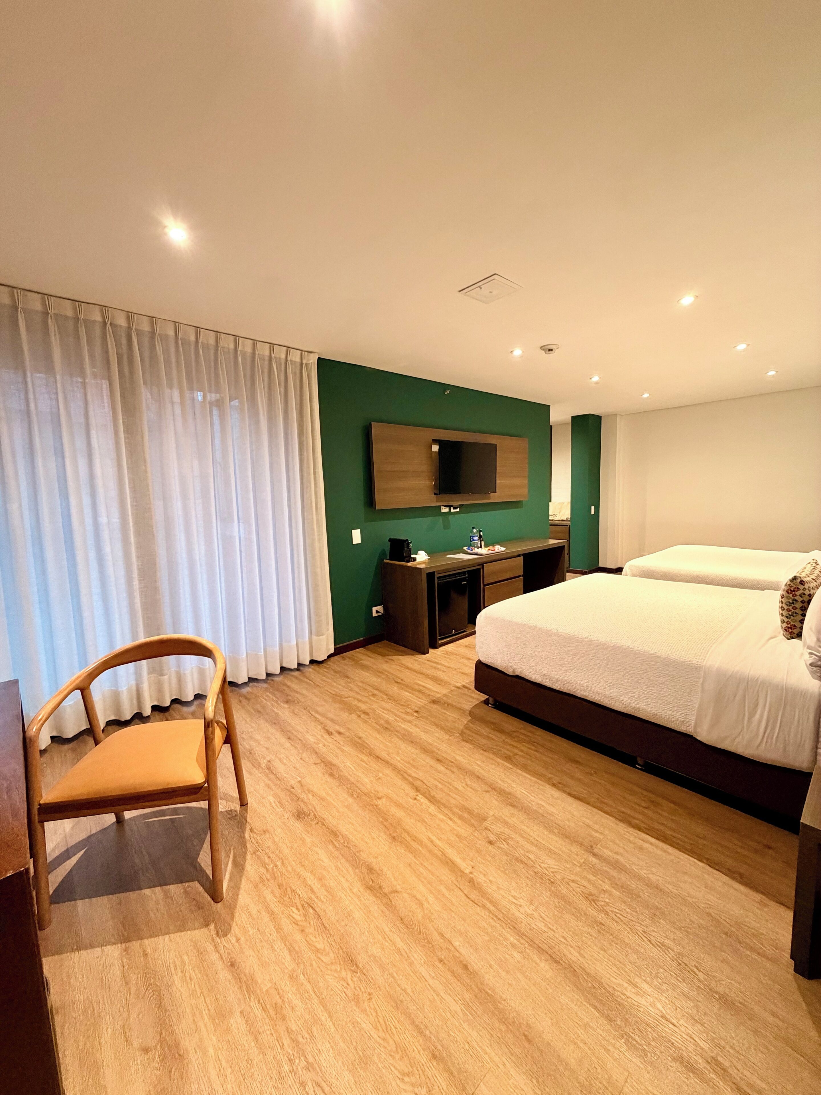 A hotel room with a large bed, a desk, a chair, a TV mounted on the wall, and a green accent wall.