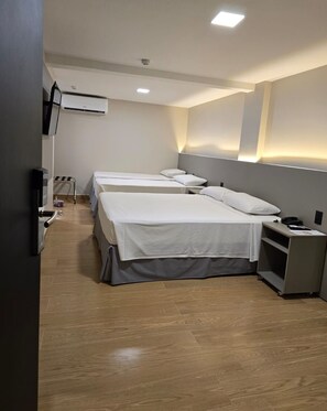 Habitación de hotel con tres camas individuales, un escritorio pequeño con una silla, un televisor de pantalla plana y un equipo de aire acondicionado instalado en la pared.