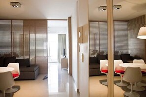 Une chambre d’hôtel moderne, équipée d’un canapé, d’une table à manger et d’une chaise. Les fenêtres sont dotées de stores, et le sol de la chambre est de couleur claire.