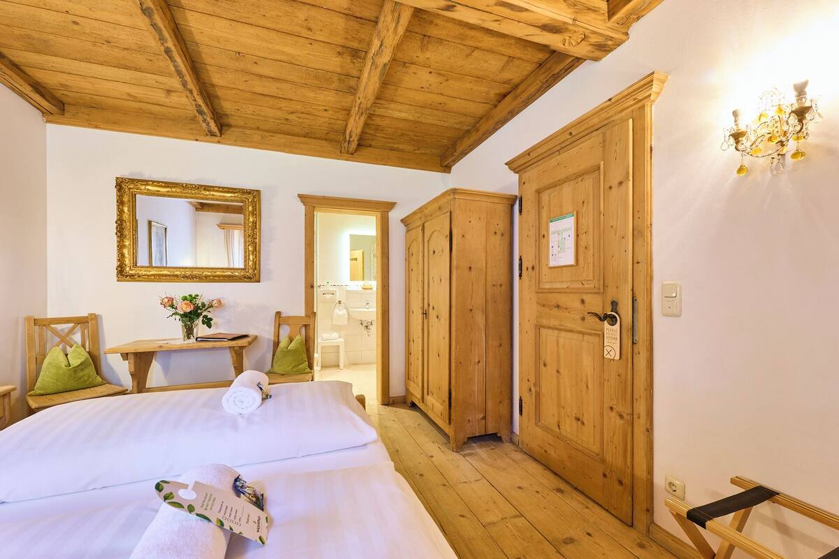 Habitación de hotel con techo de madera, cama con ropa de cama blanca, armario de madera, pequeña mesa con un jarrón de flores y espejo con marco dorado.