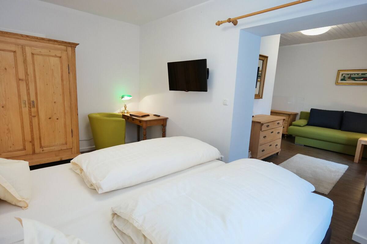 Habitación de hotel con dos camas, un escritorio con lámpara, una silla verde, un armario de madera, un sofá verde y un televisor en la pared.