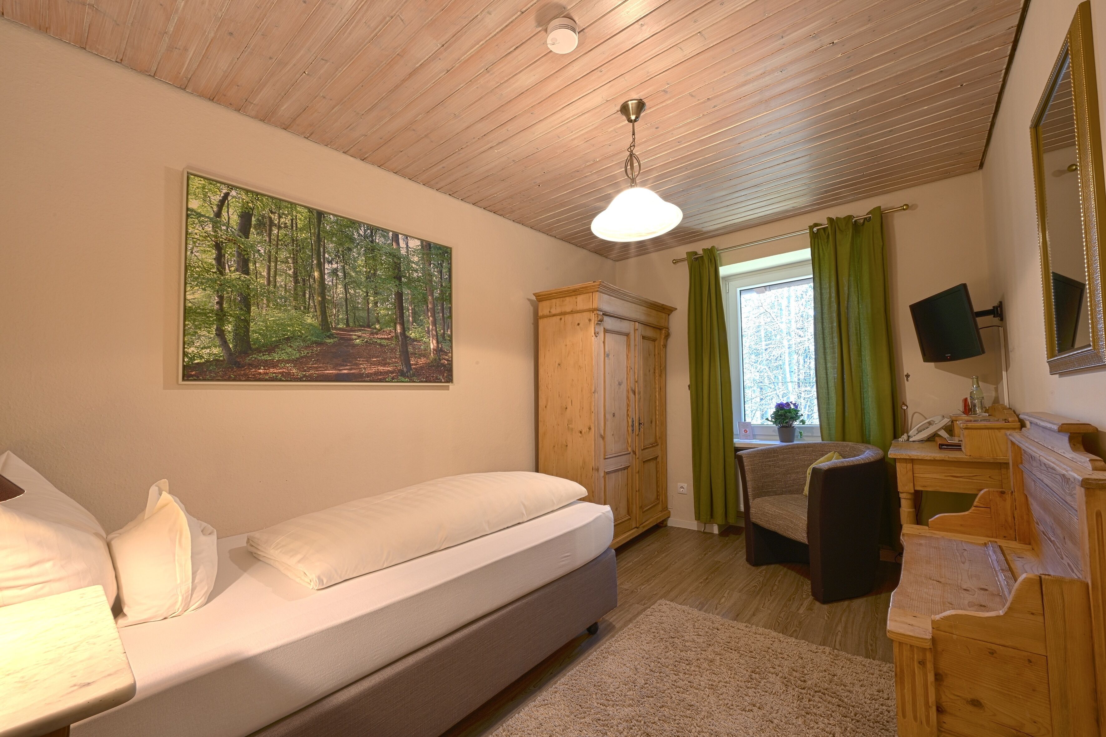 A bedroom with a bed, a wooden wardrobe, a desk with a chair, and a painting of a forest on the wall.