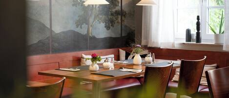 A restaurant interior with wooden benches, a table set for two, and a large window with a view of greenery.