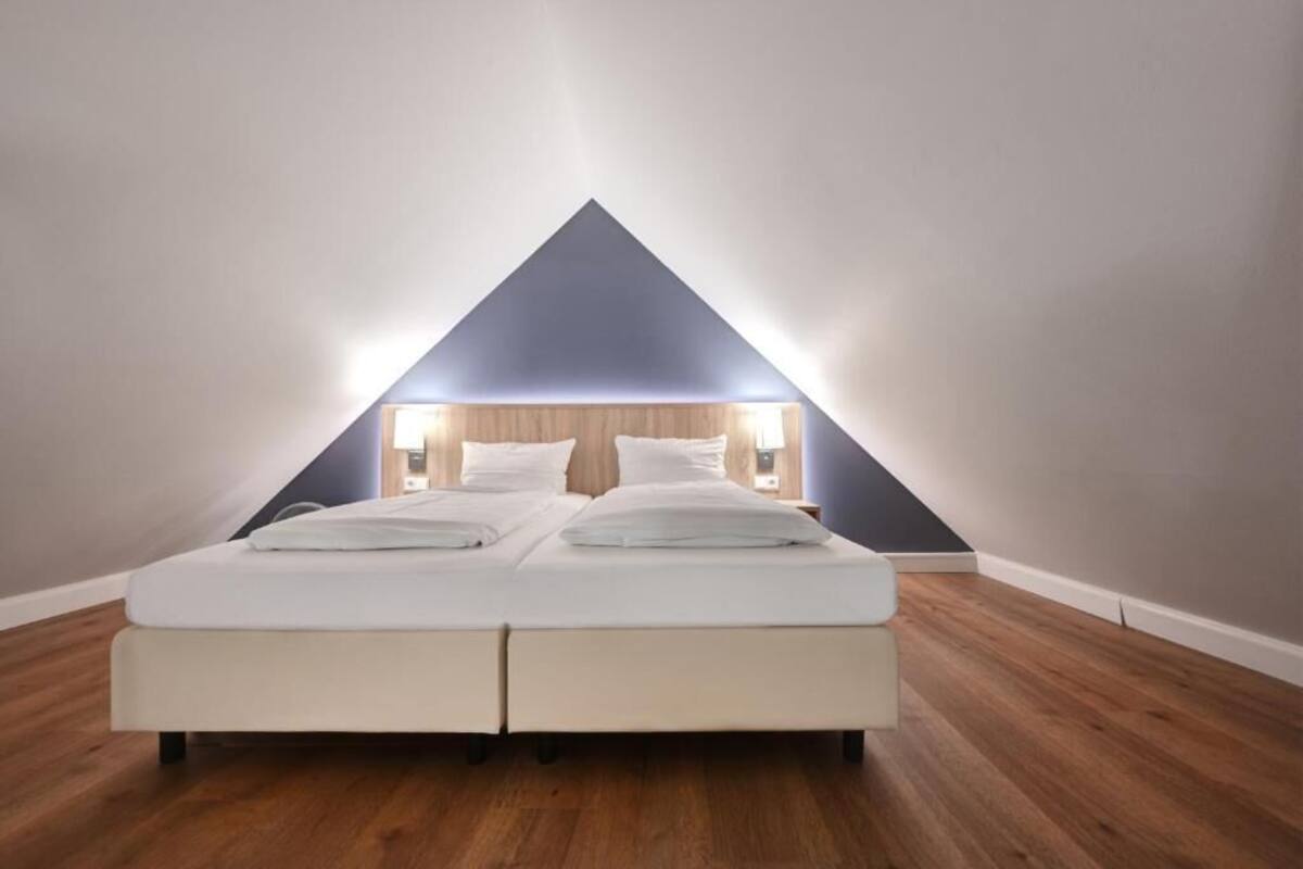 Camera da letto con un letto al centro sotto un soffitto triangolare, pavimento in legno e un arredamento semplice e moderno.