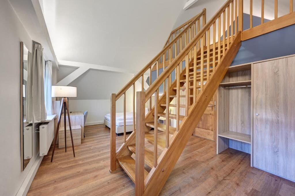 A wooden staircase leading to a loft bedroom with a sloped ceiling, a desk with a lamp, and a bed.
