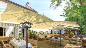 An outdoor patio with wooden tables and benches, covered by large umbrellas, and a brick floor. - Weichandhof (Munich)