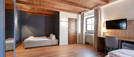 A hotel room with wooden flooring, a bed, a desk, a chair, a television, and a window.
