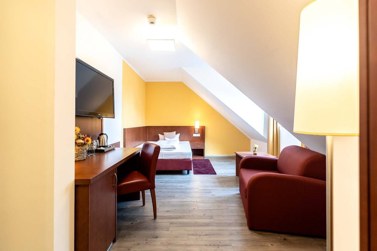 Una camera d'albergo con una scrivania, un letto, un divano rosso e una televisione.