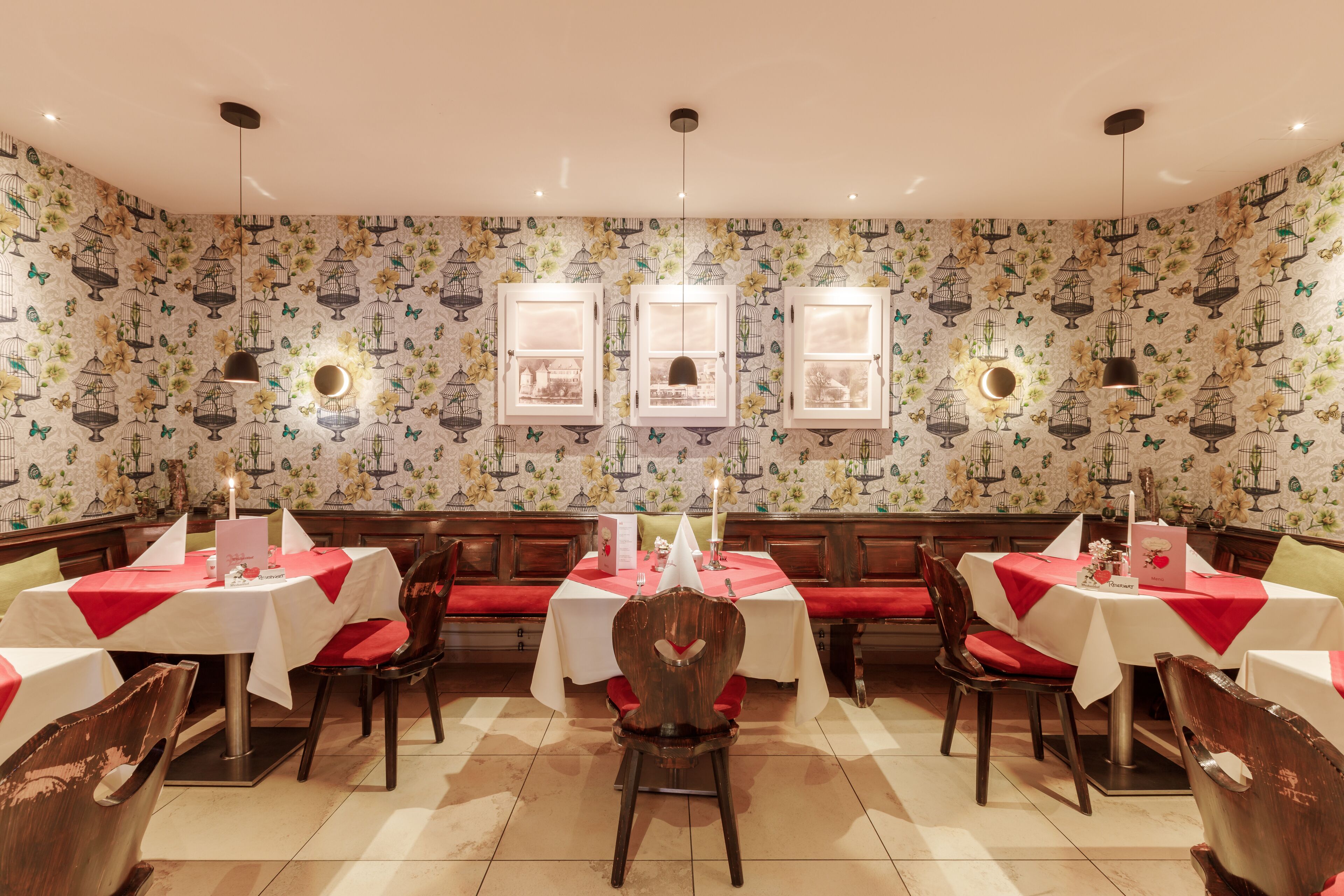 Un restaurante con decoración clásica, que presenta mobiliario de madera, manteles blancos y rojos, y papel tapiz con motivos de pájaros.
