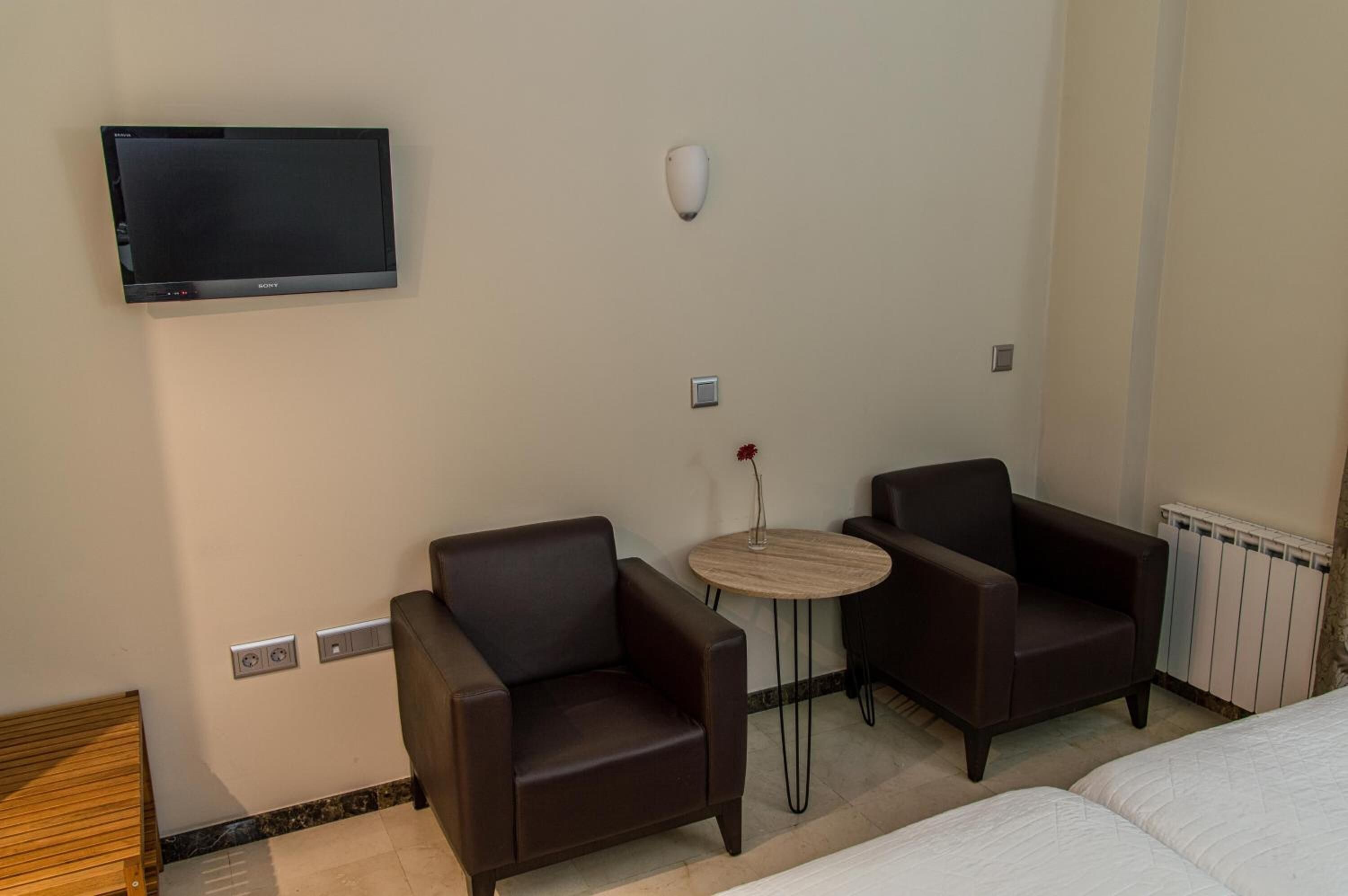 A hotel room with a bed, two armchairs, a small round table, a wall-mounted TV, and a radiator.
