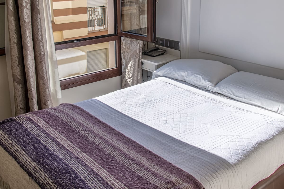 A neatly made bed with a striped blanket, a bedside table with a lamp, and a window with curtains.