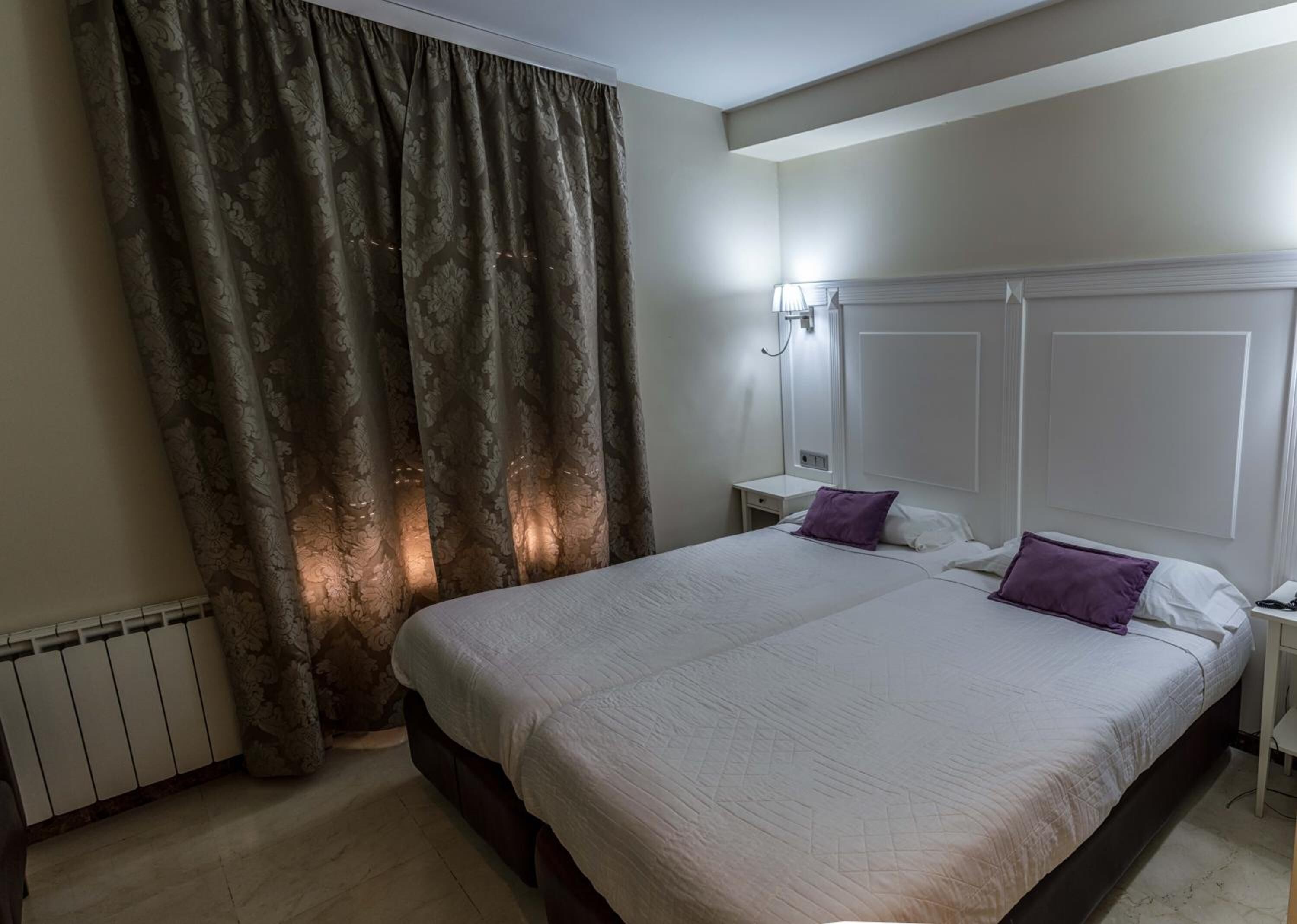 Quarto de hotel com uma cama grande, duas mesas de cabeceira, um criado-mudo, uma luminária e uma janela com cortinas.