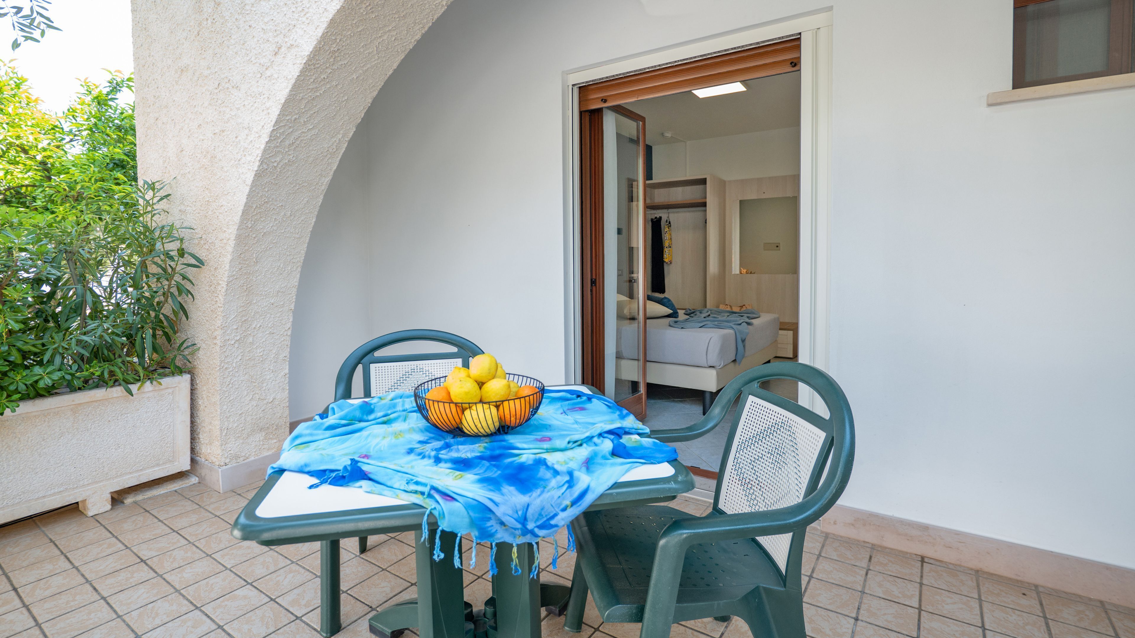 A small patio with a table and chairs, a bowl of fruit, and a view into a bedroom.