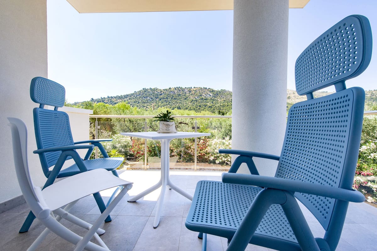 A balcony with two blue chairs and a small table, overlooking a green landscape with hills.
