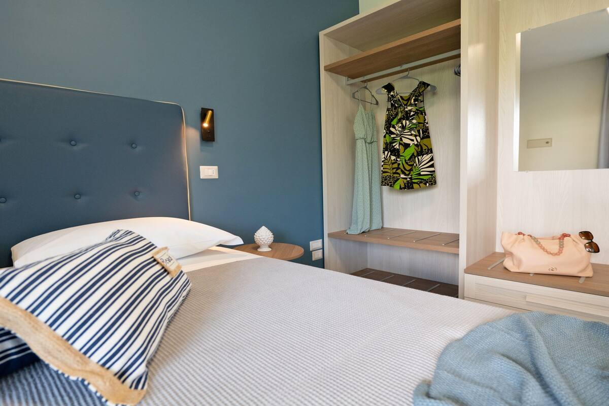 A bedroom with a tufted headboard, a bed with striped pillows, a bedside table with a lamp, and a bench with a dress and a handbag.