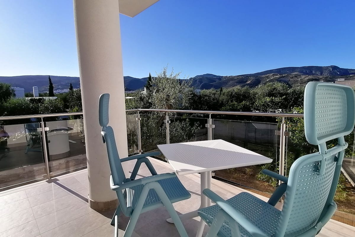 A balcony with two blue chairs and a white table, overlooking a scenic landscape with mountains and trees.