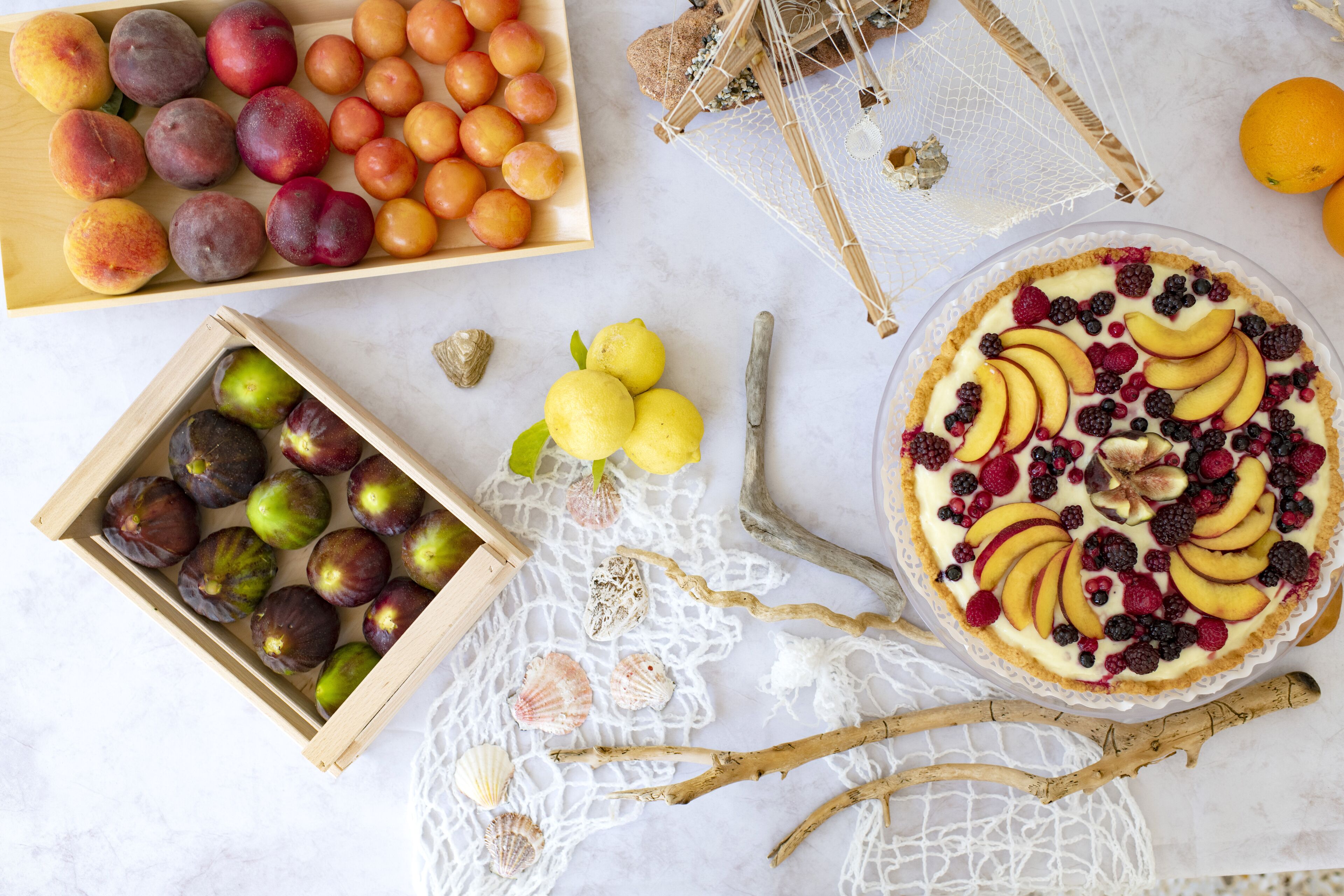 A wooden crate with various fruits, a basket of plums, and a pie with fruit toppings.