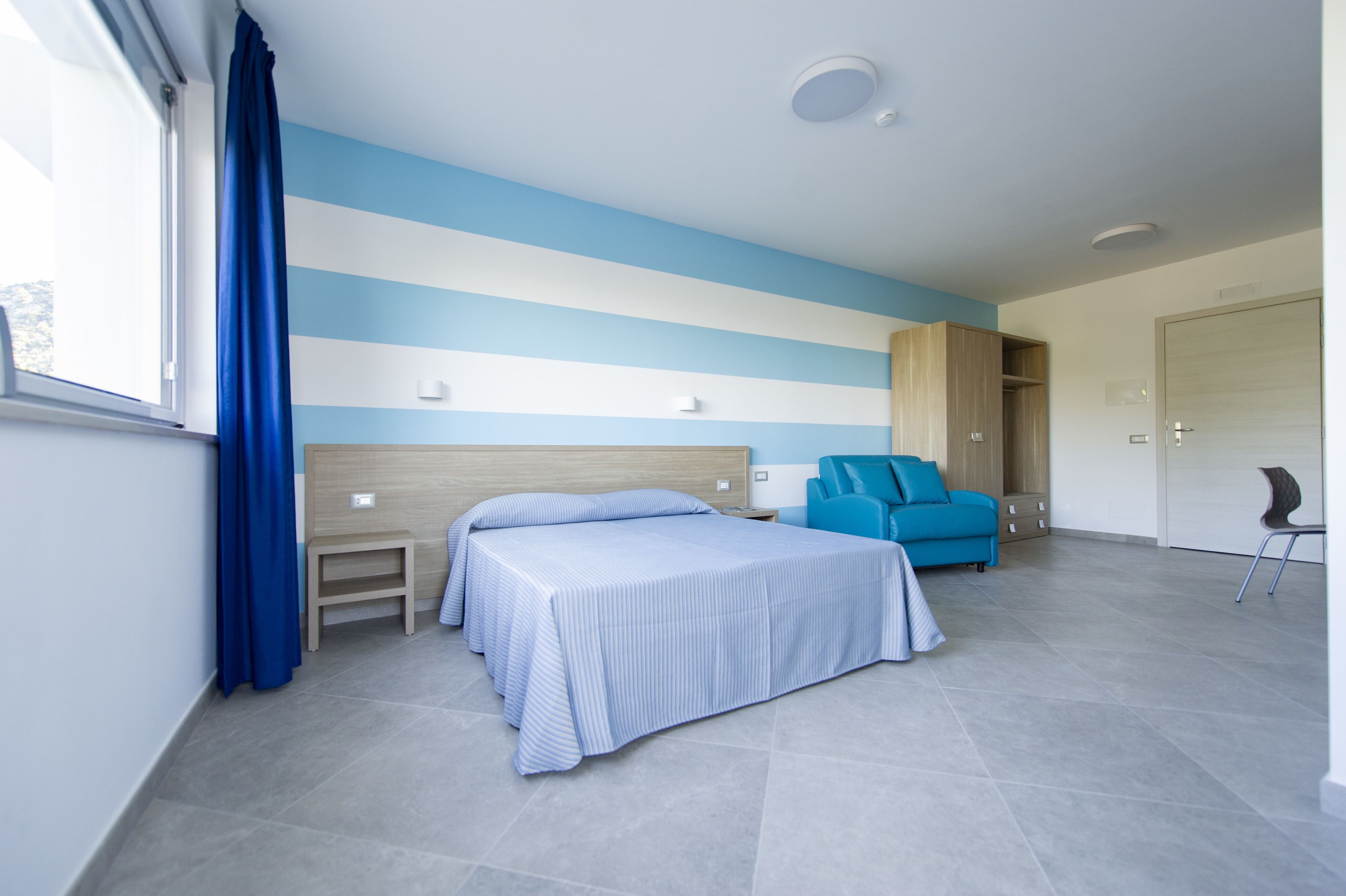 A bedroom with two beds, a blue chair, a wardrobe, and a small table.