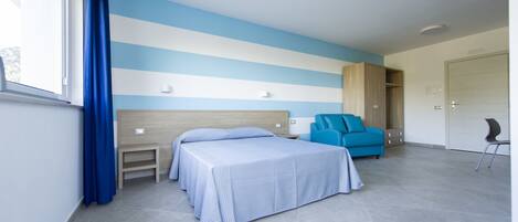 A bedroom with two beds, a blue chair, a wardrobe, and a small table.