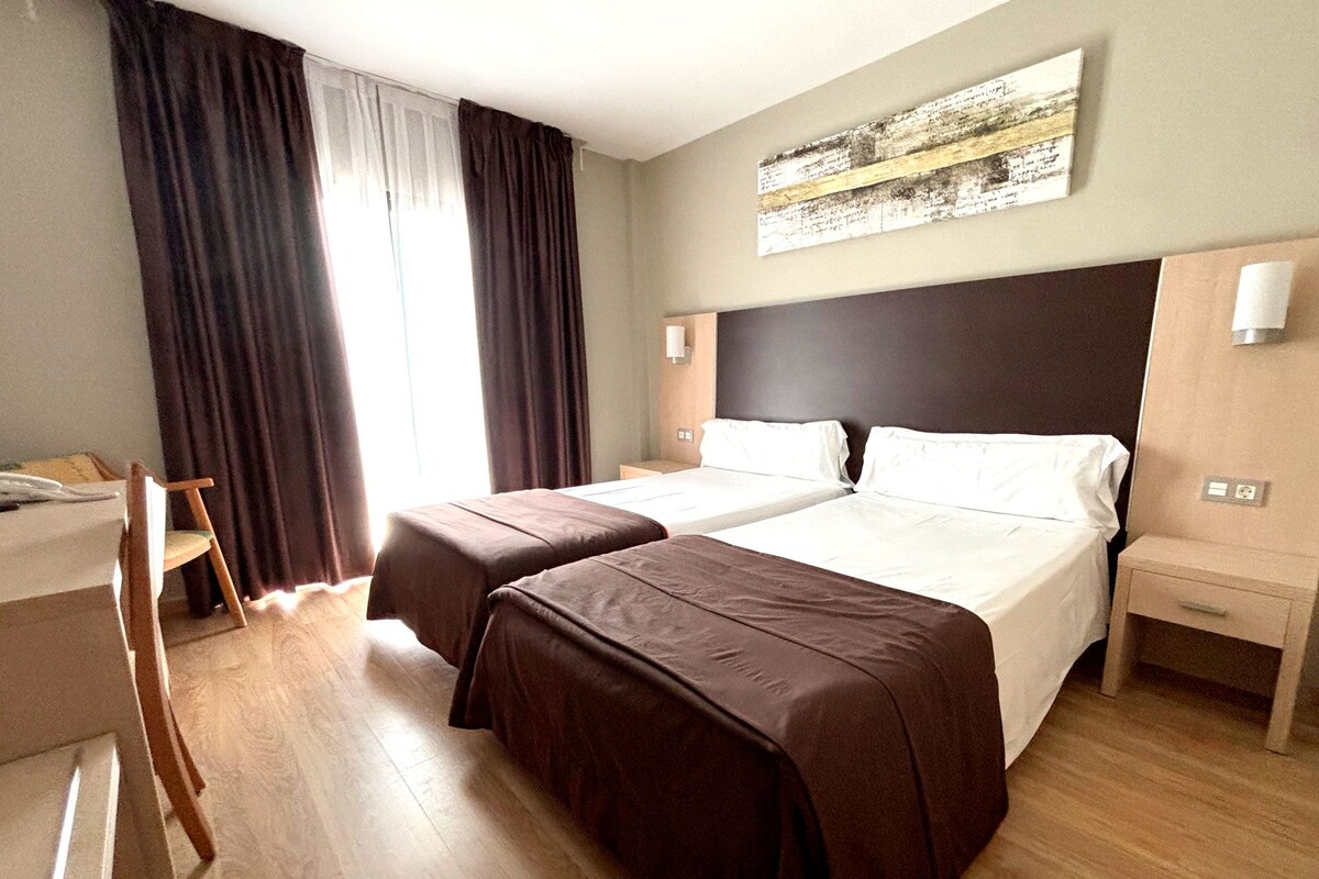 A hotel room with two beds, a desk, a chair, and a large window with curtains.