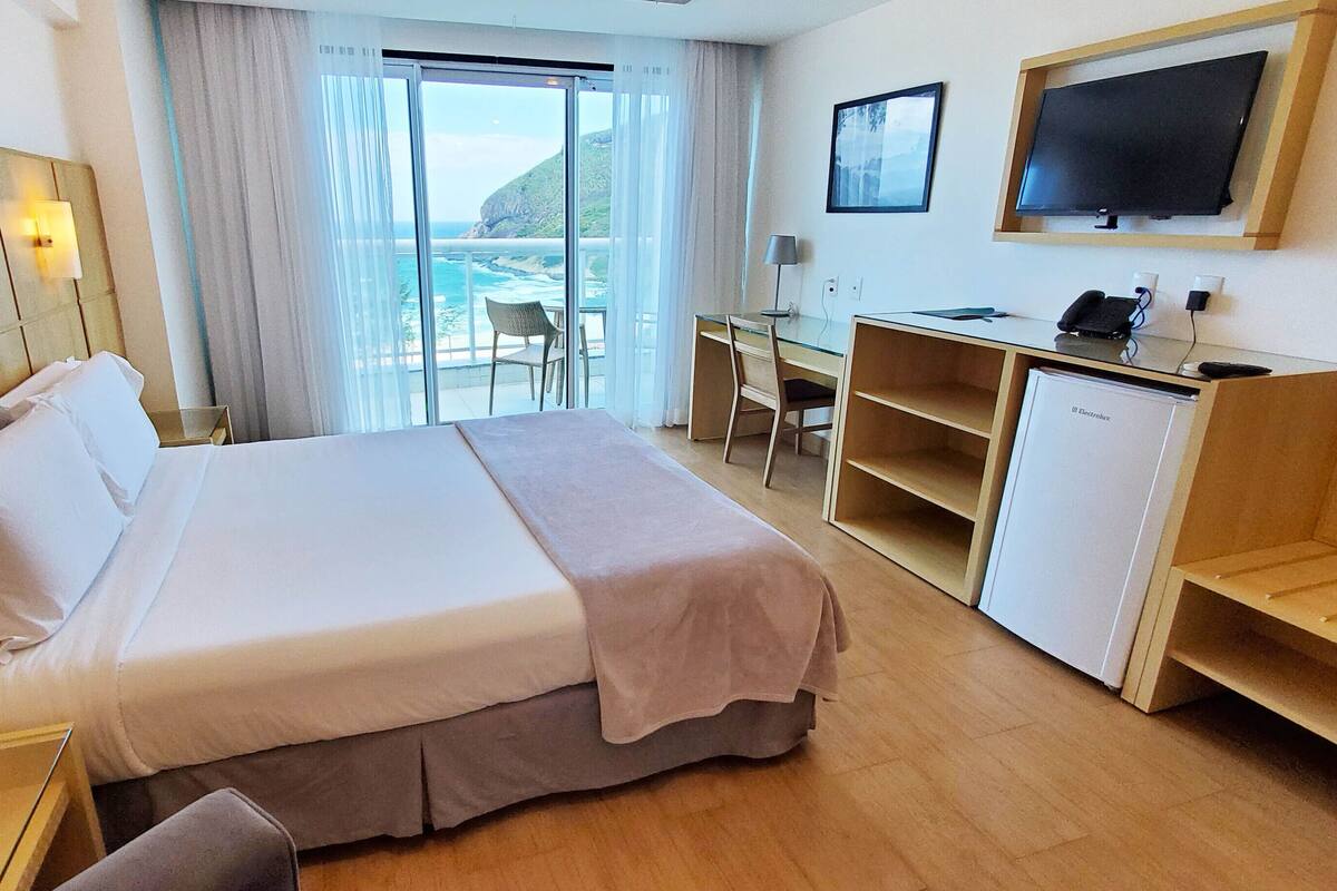 Een hotelkamer met een bed, een bureau, een stoel, een televisie en uitzicht op de oceaan.