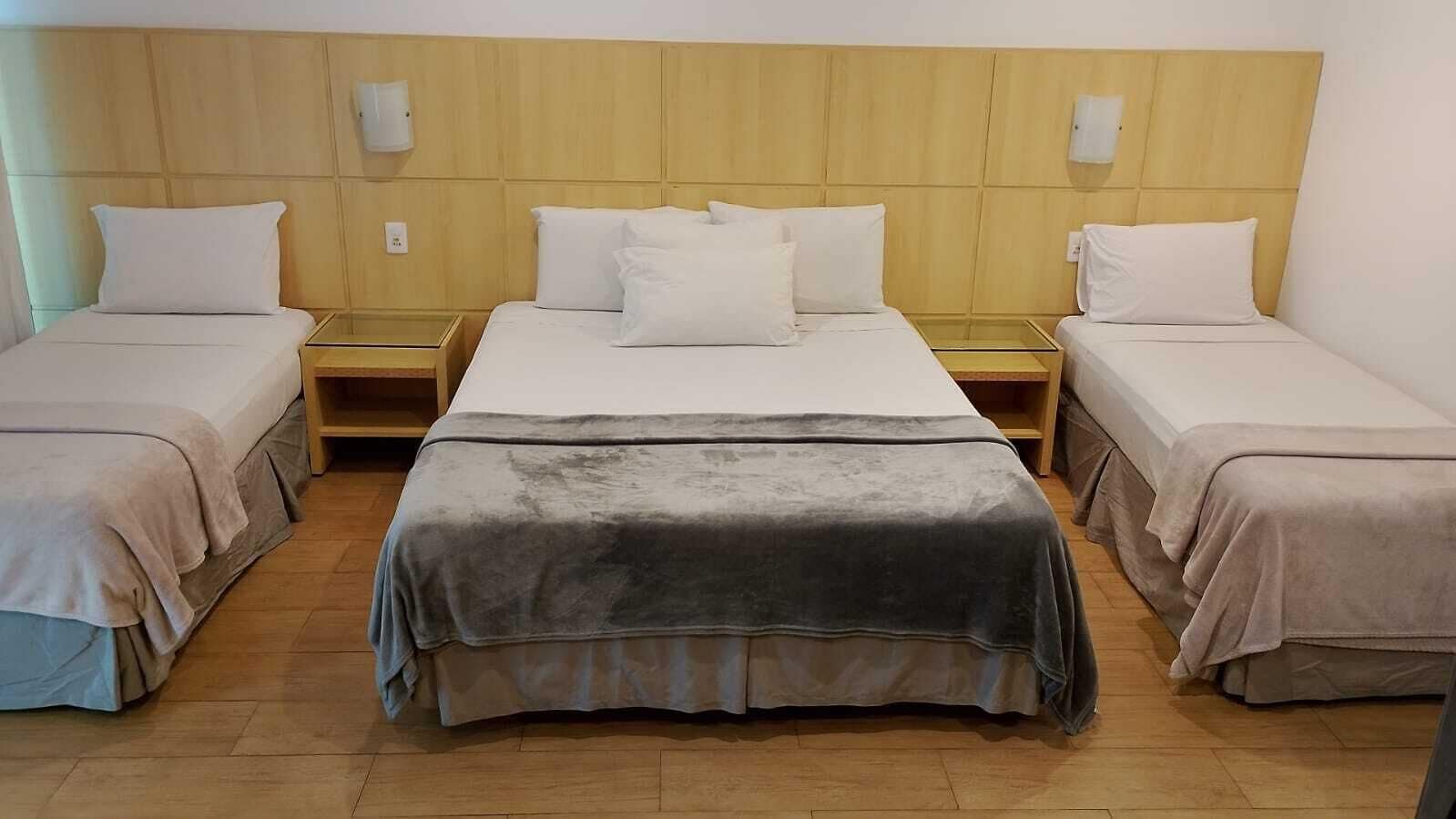 Egy szállodai szoba, amelyben egy nagy ágy, két kisebb ágy, fafejtámlás ágykeret, éjjeliszekrények és egy szürke takaró található.