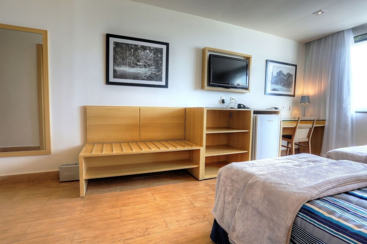 A hotel room with a wooden bench, a desk, a bed, and two framed pictures on the wall.