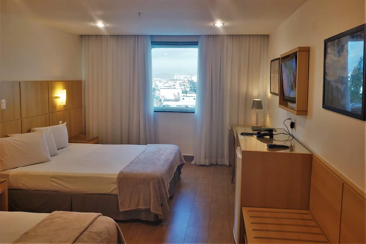 Een hotelkamer met een bed, een bureau, een stoel, een raam met gordijnen en uitzicht over de stad.