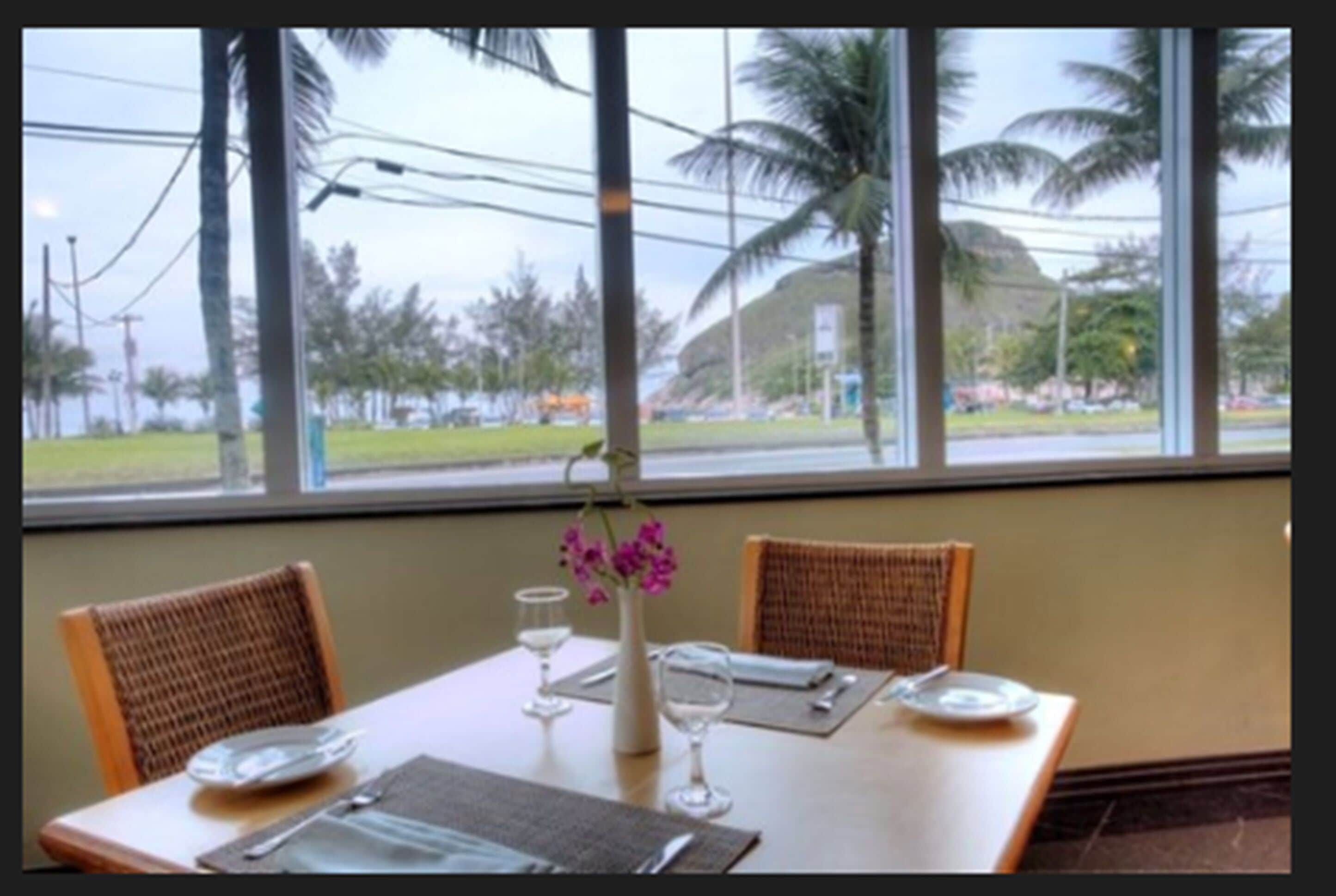 A dining table with chairs, a vase with flowers, and a view of palm trees and a building through large windows.