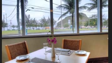 A dining table with chairs, a vase with flowers, and a view of palm trees and a building through large windows.