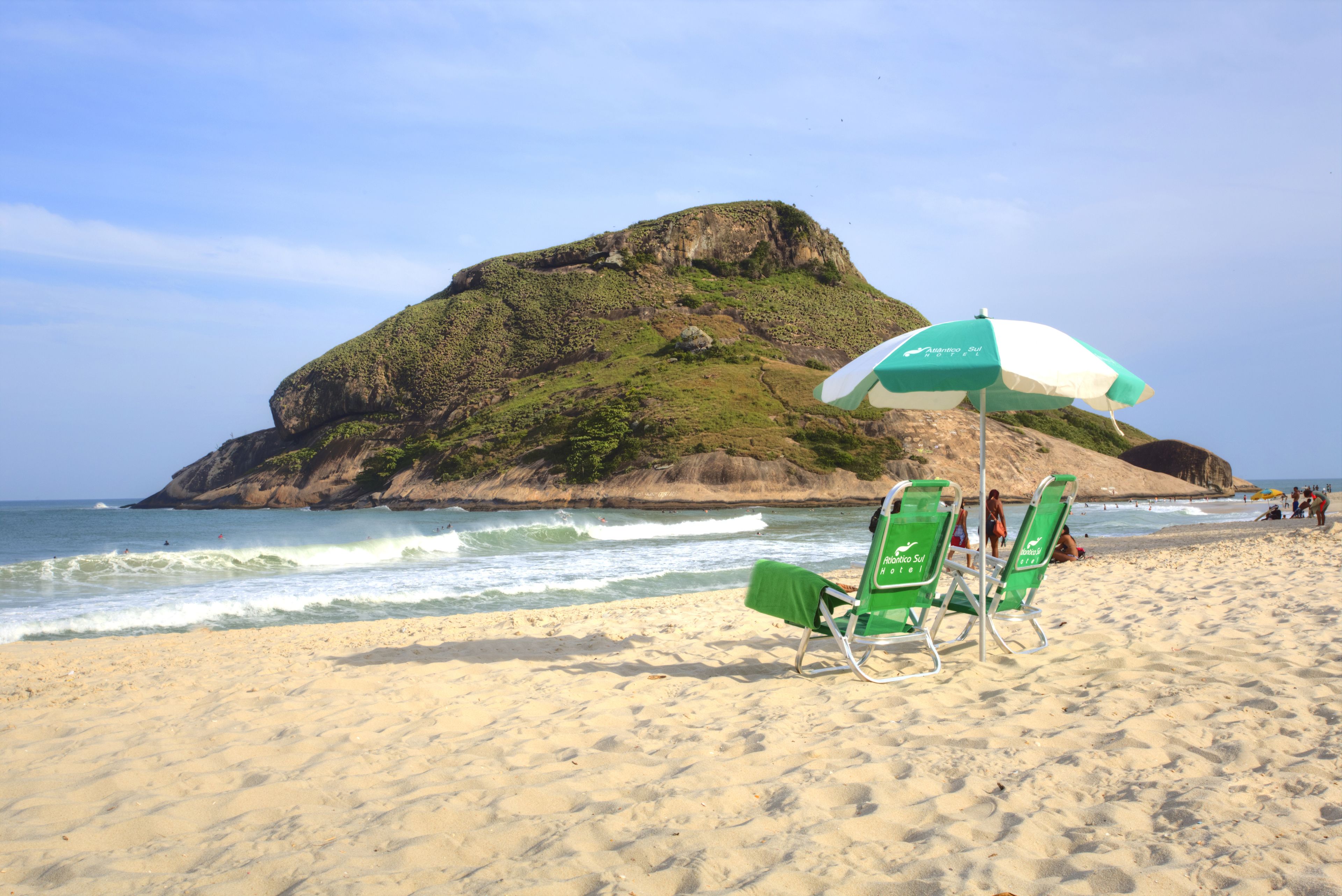 Una spiaggia con due poltrone verdi sotto un ombrellone bianco e verde, una collinetta rocciosa sullo sfondo e l'oceano.