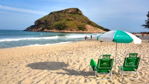 Een strand met twee groene ligbedden onder een witgroene parasol, zandstrand en een rotsachtige heuvel op de achtergrond.