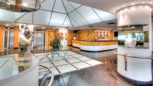 Een moderne lobby met een glazen receptiebalie, een centrale zithoek en een hoog plafond met geometrische patronen.