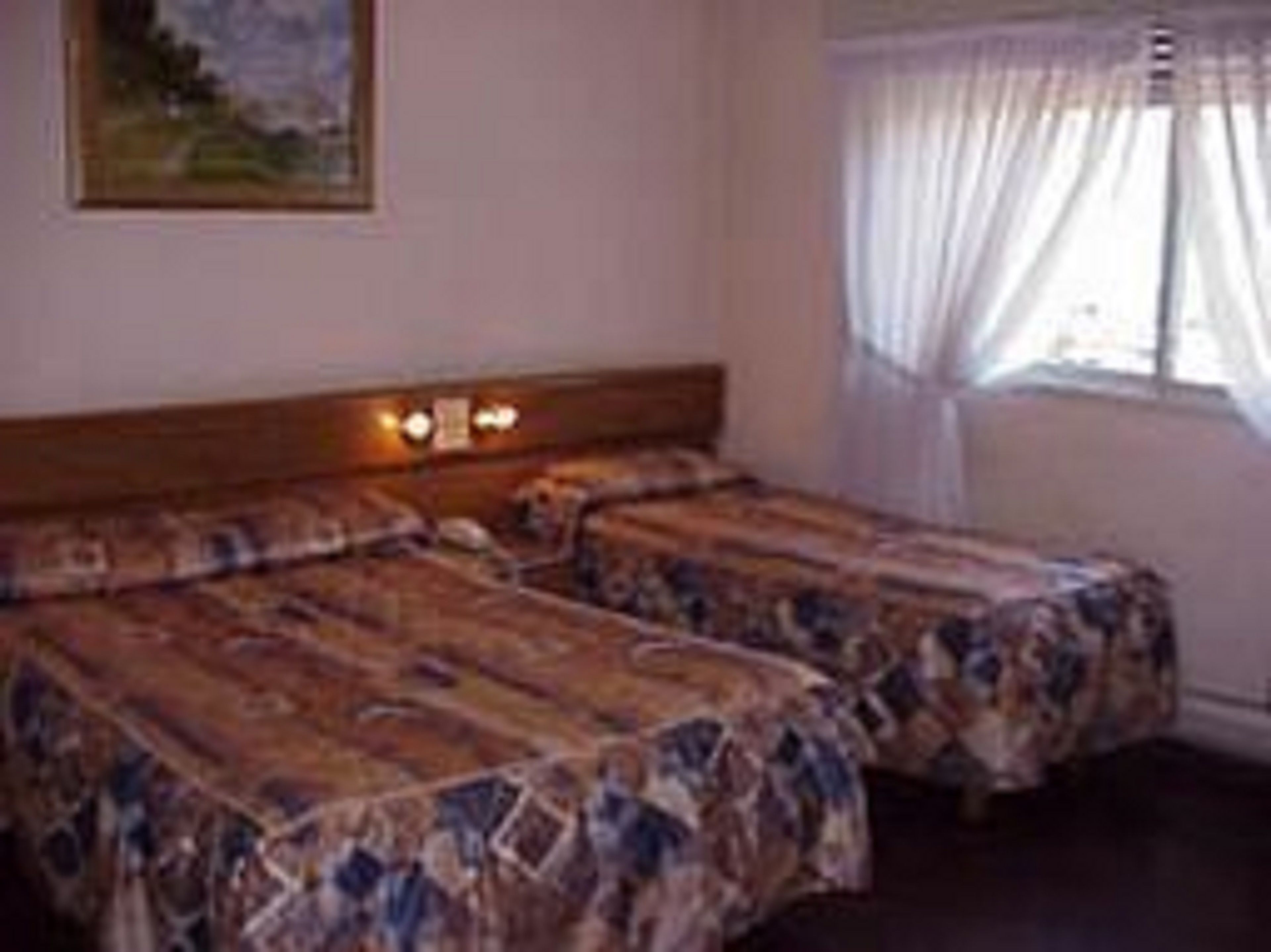 A room with two single beds, a wooden headboard, a painting on the wall, and a window with sheer curtains.