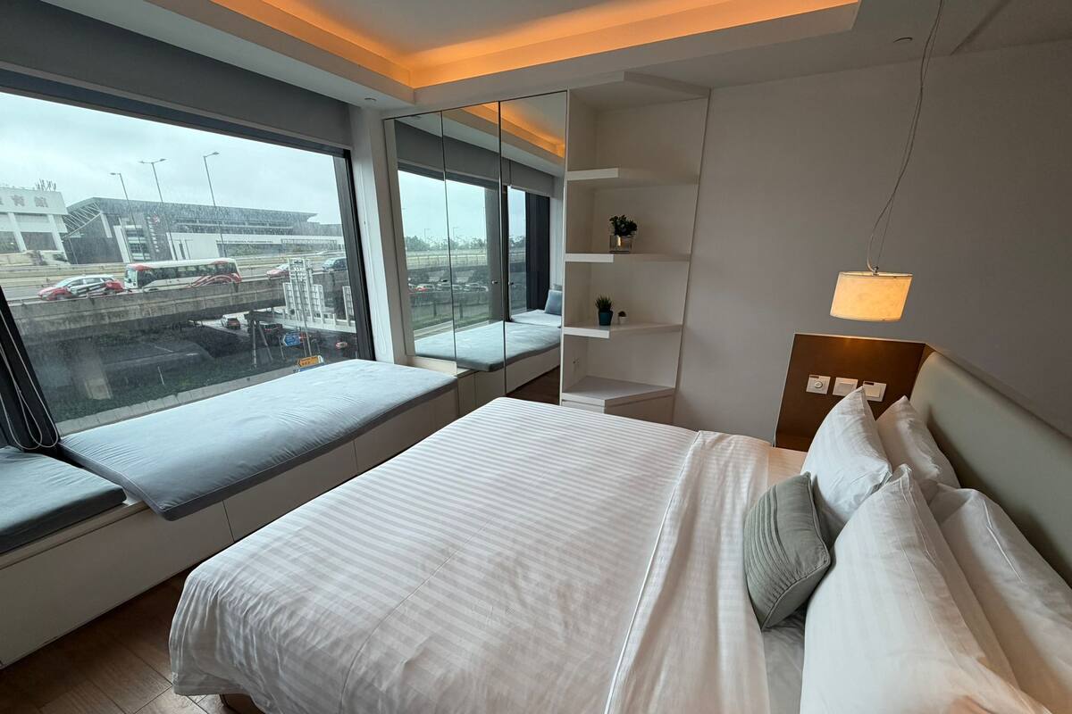 A modern hotel room with a large bed, a bedside table with a lamp, a small shelf, and a window seat overlooking a busy street.