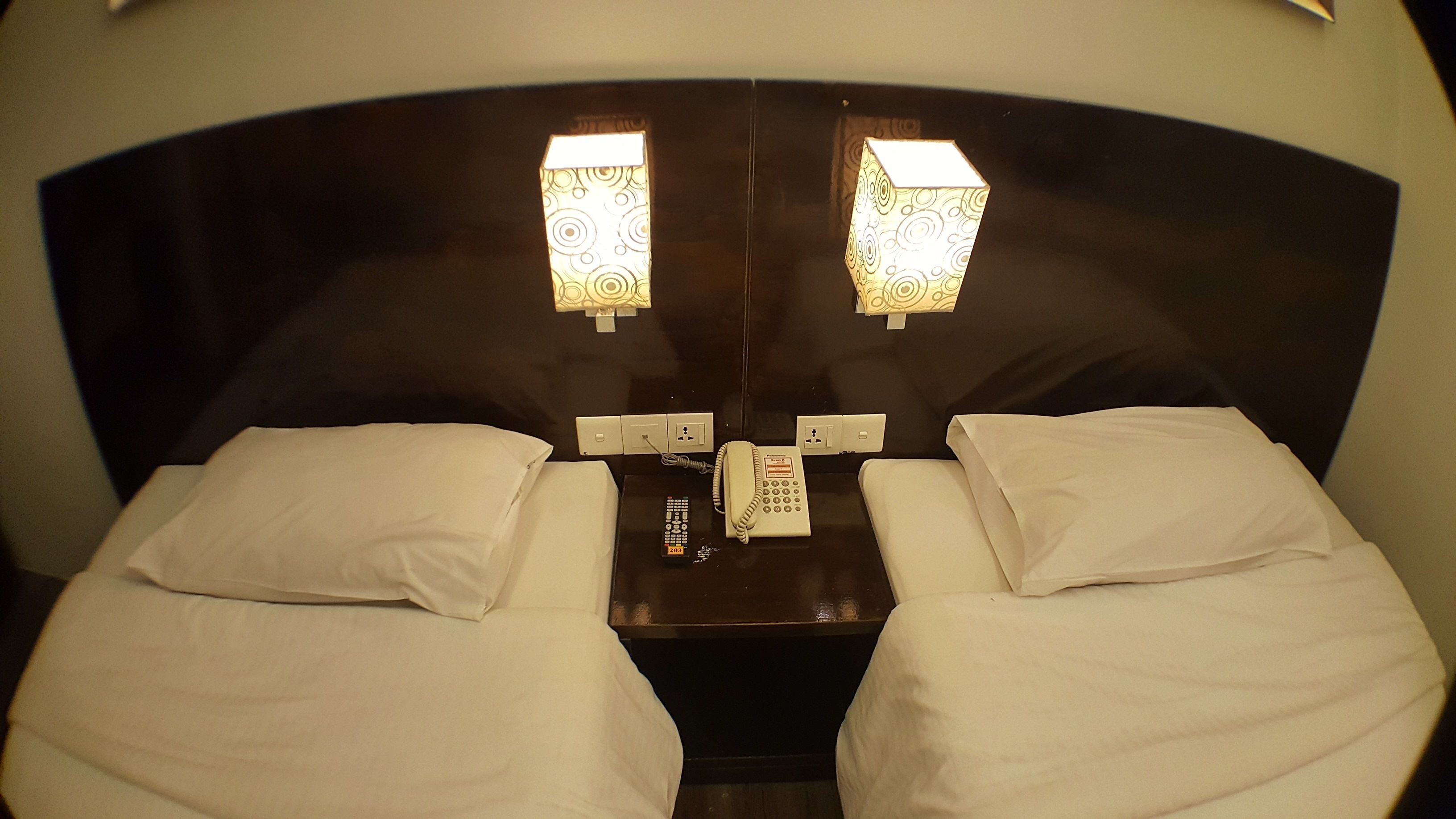 A hotel room with two beds, a nightstand with a telephone and remote control, and wall-mounted light fixtures.