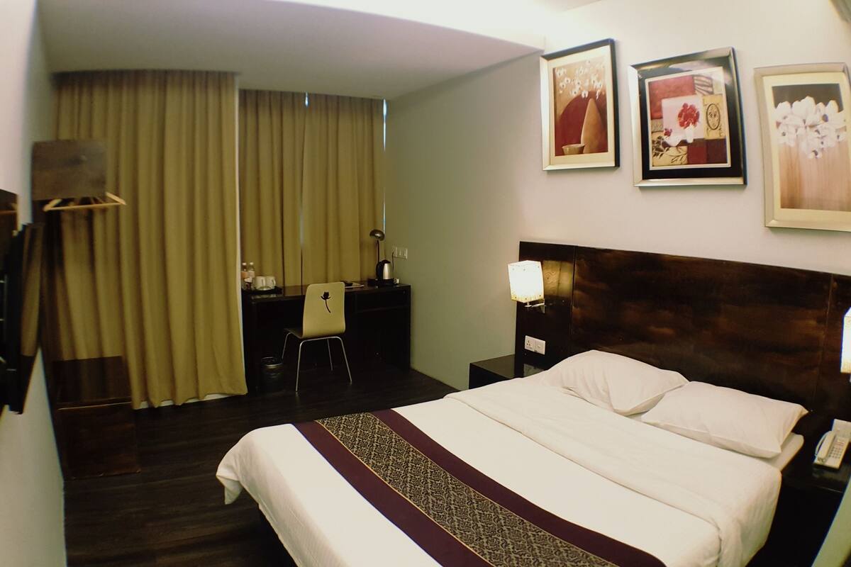 A hotel room with a bed, a desk, a chair, a desk lamp, a telephone, and a wall-mounted TV.