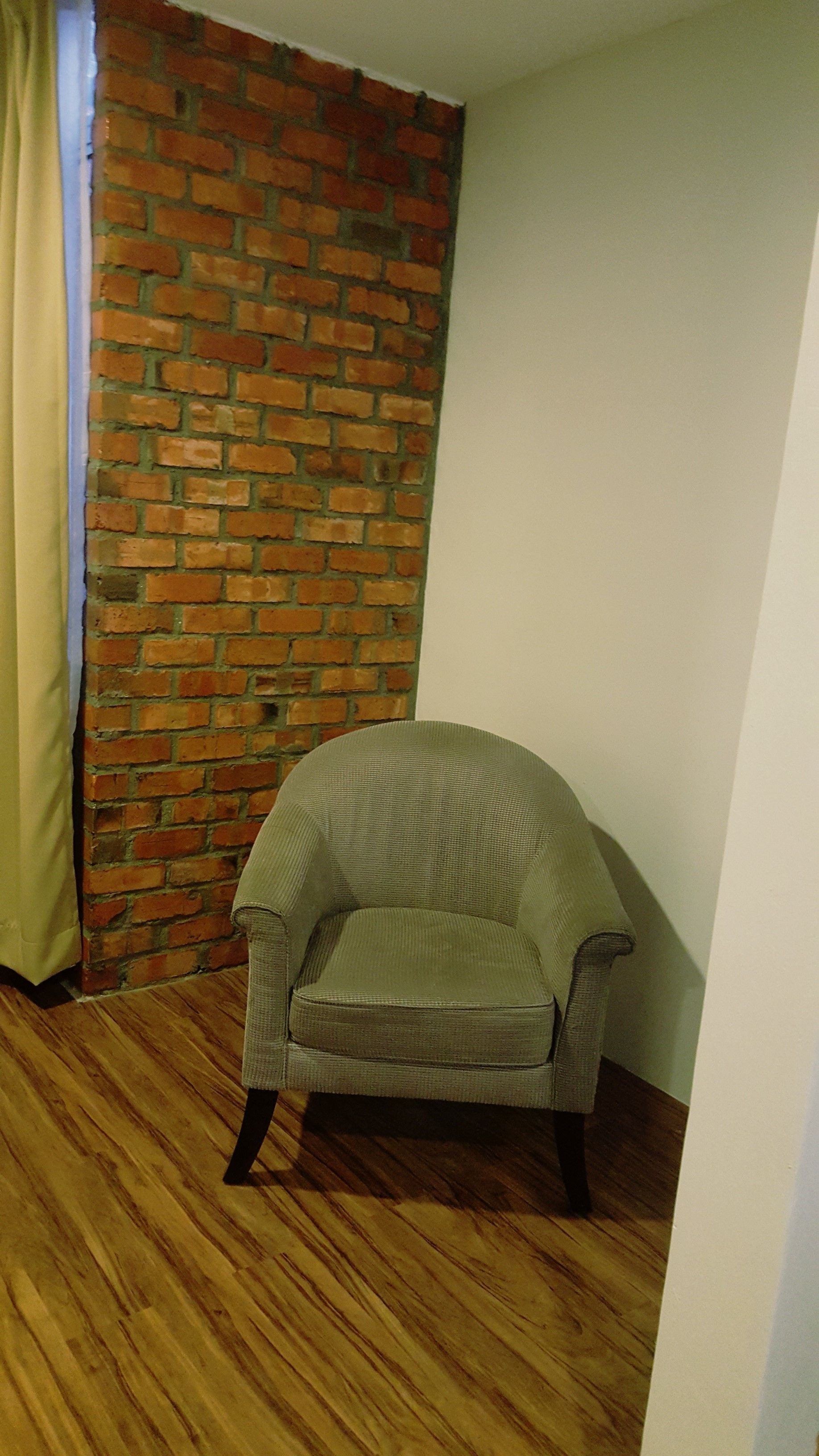 A corner room with a brick wall, a wooden floor, and a single armchair.