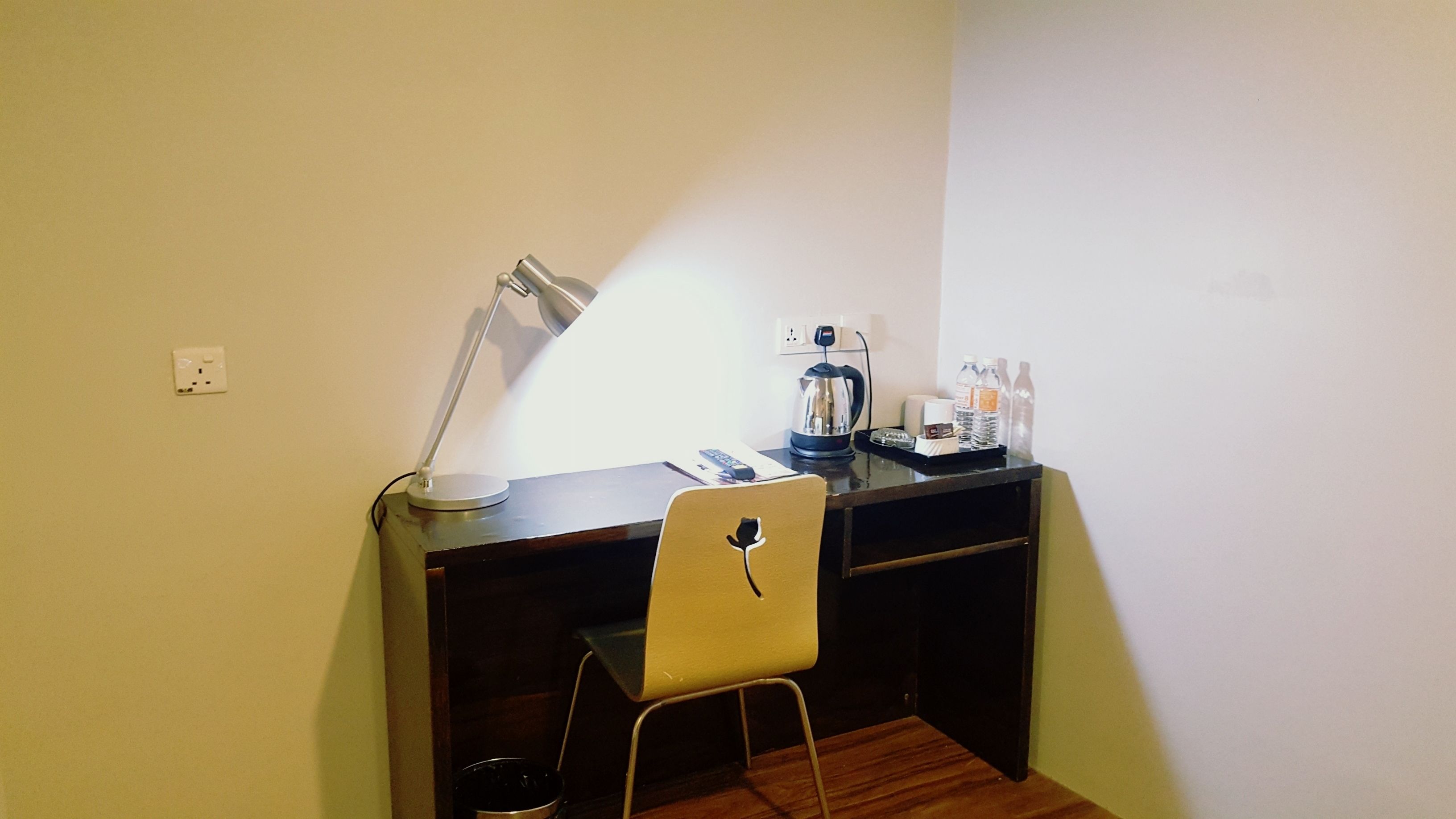 Una habitación pequeña y bien iluminada con un escritorio, una silla y una lámpara. Hay un hervidor, un control remoto y algunas botellas sobre el escritorio.