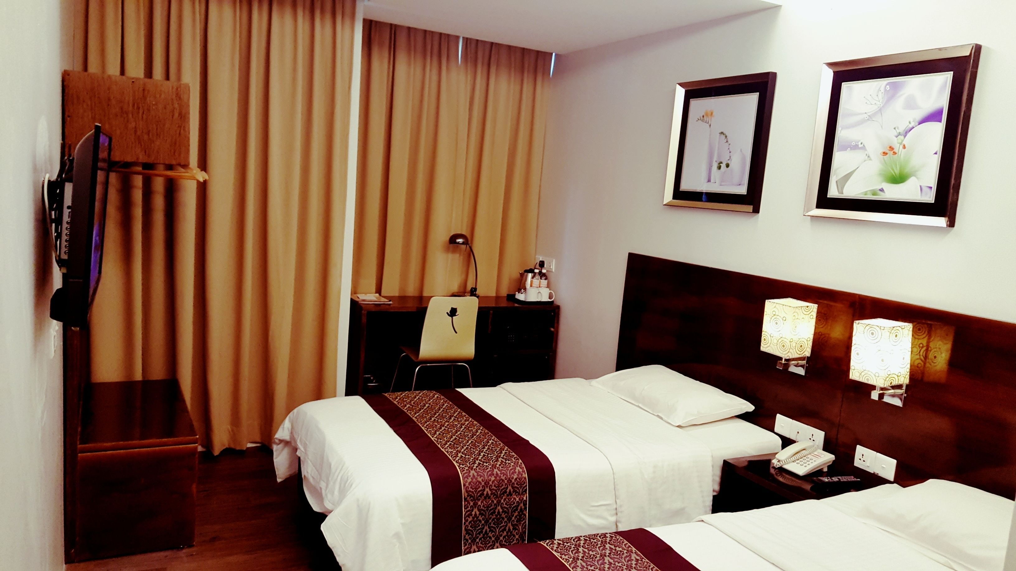 A hotel room with two beds, a desk with a chair, a TV, and two framed pictures on the wall.