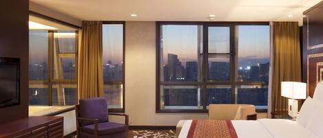 A hotel room with a large bed, a chair, a TV, and a view of the cityscape through large windows.