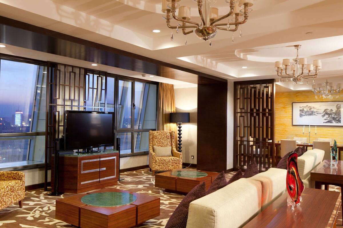 A modern hotel room with a large TV, comfortable seating, a dining area, and a view of the cityscape.