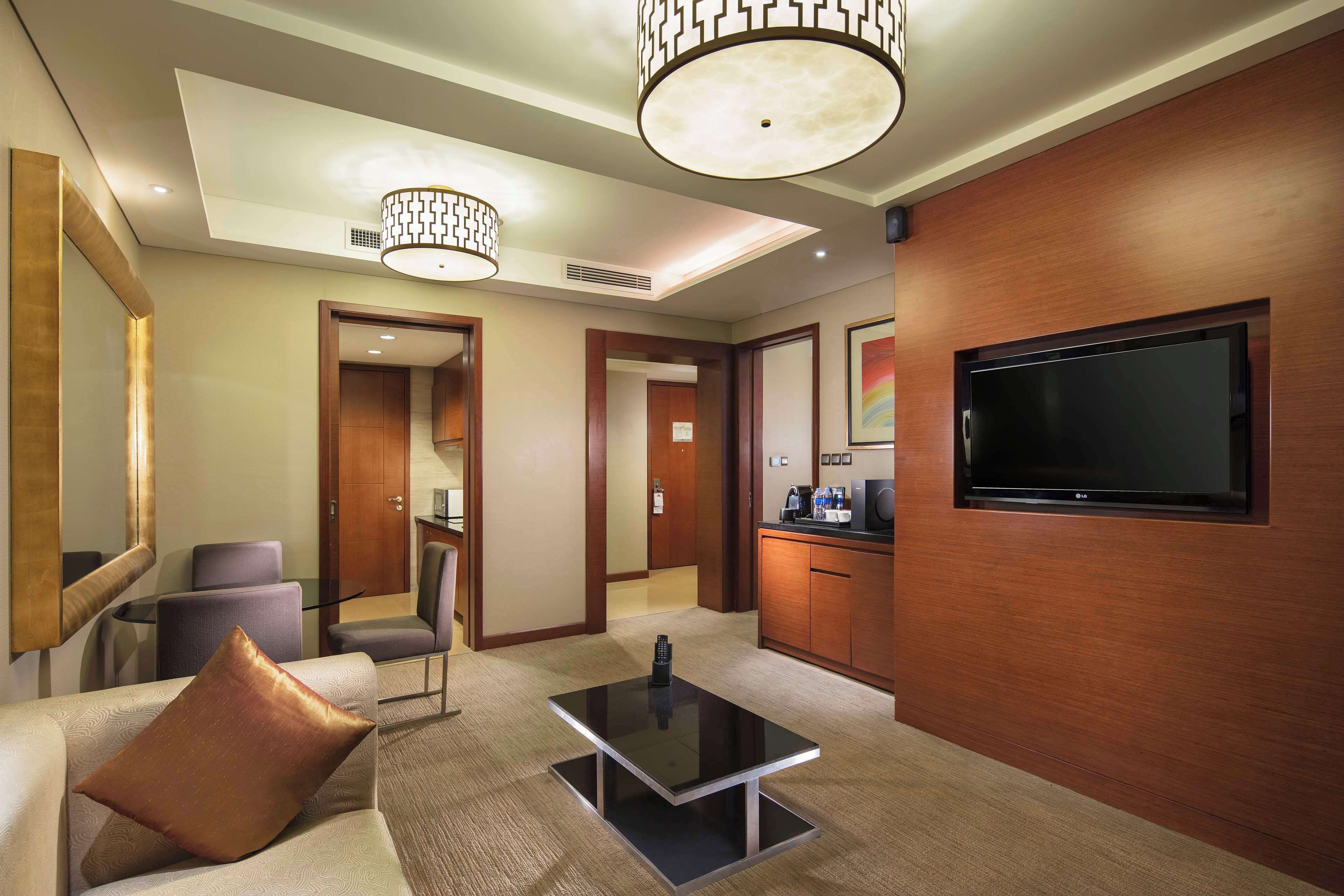 A hotel room with a flat-screen TV mounted on the wall, a sofa with a decorative pillow, a glass coffee table, and a modern ceiling design with pendant lighting.