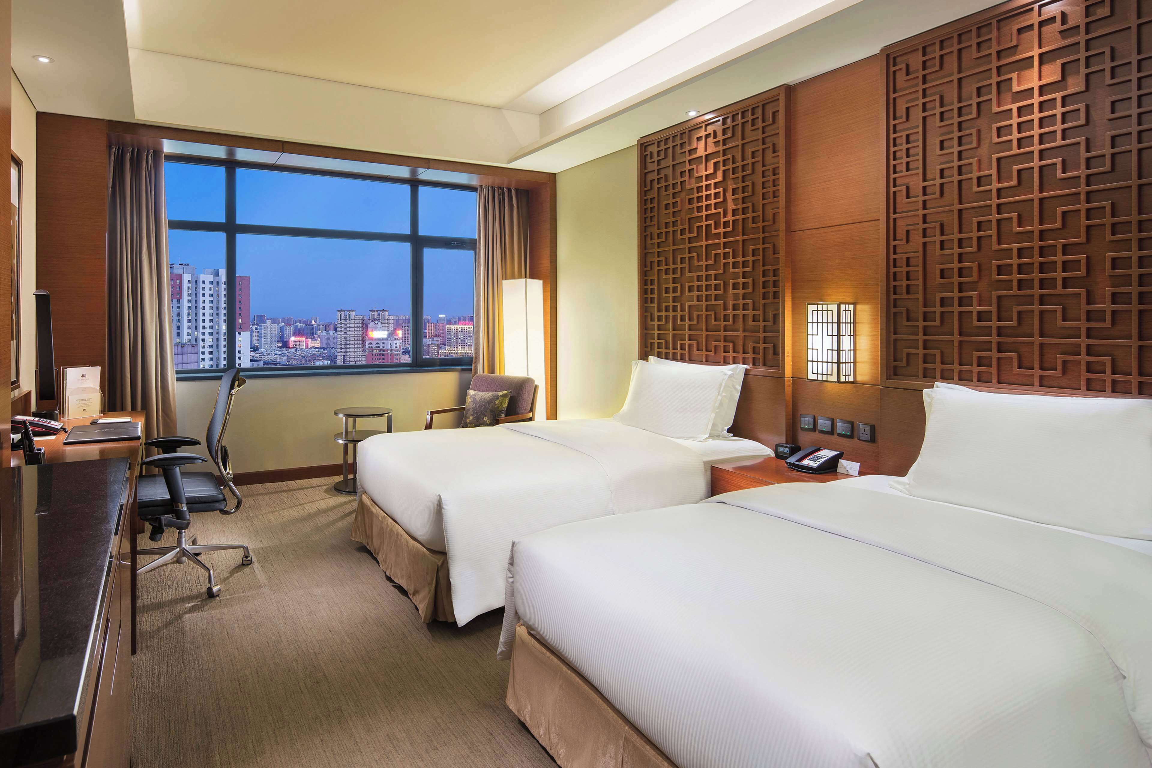 A hotel room with two beds, a desk, a chair, a lamp, and a view of the cityscape.