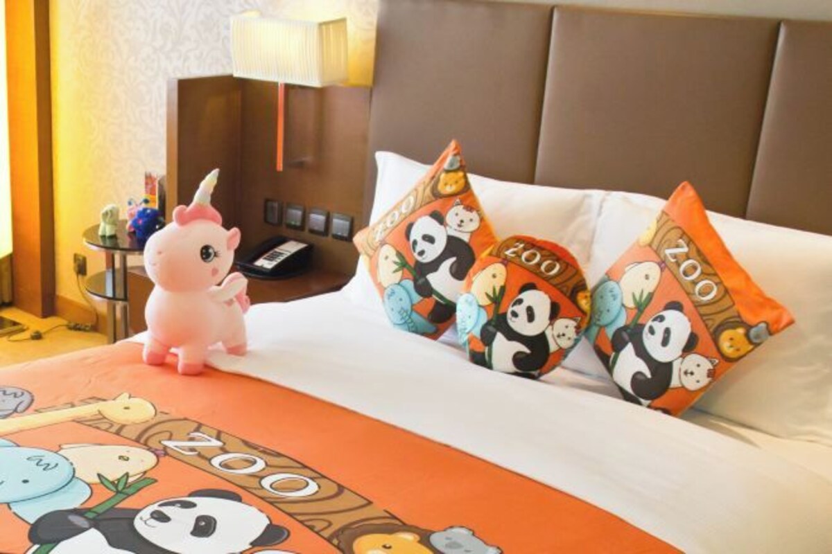 Una habitación de hotel con cama, almohadones con diseño de panda, una pequeña figura de unicornio rosa y una colcha con motivos de panda.