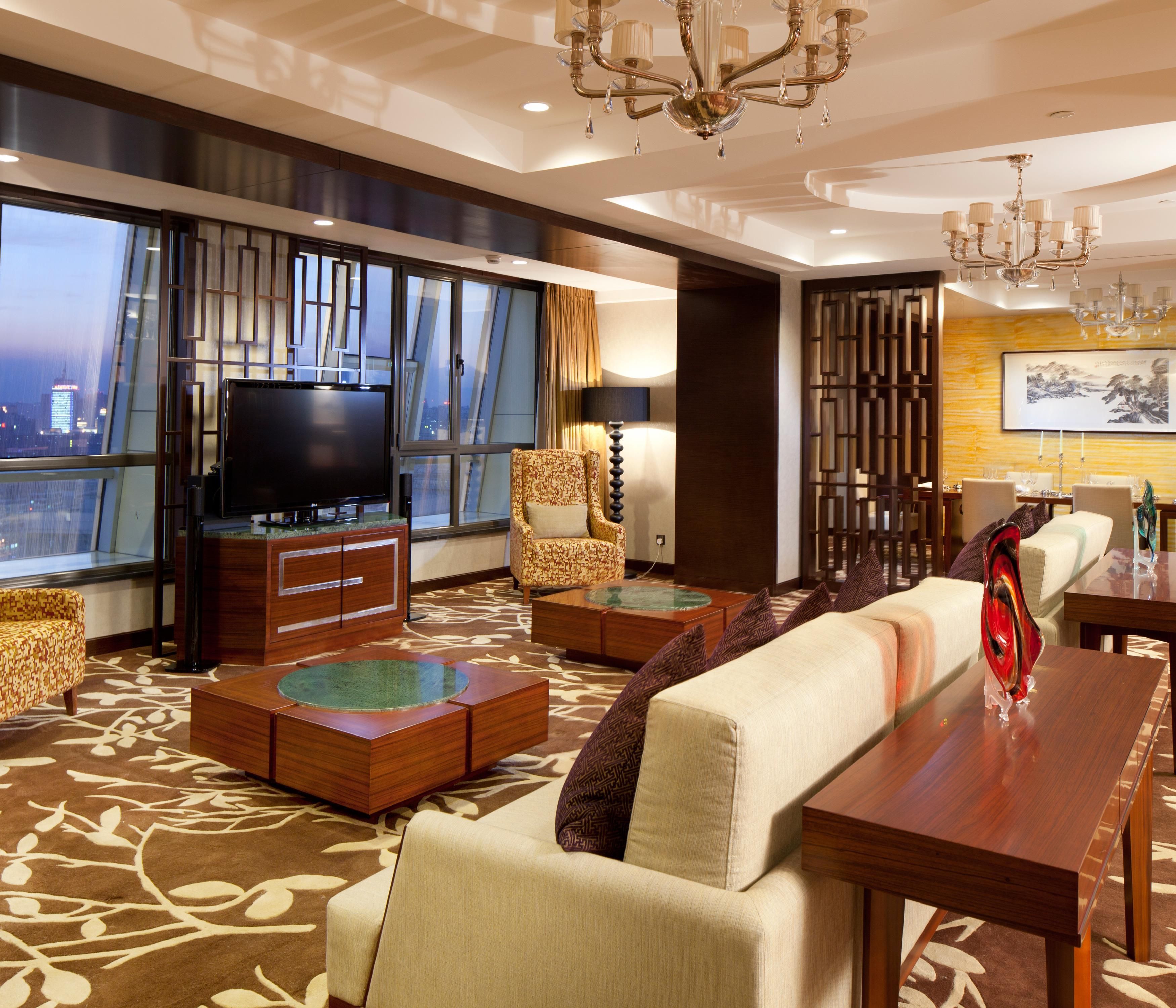 Habitación de hotel moderna con una televisión grande, asientos cómodos, una mesa de comedor y vistas a la ciudad.