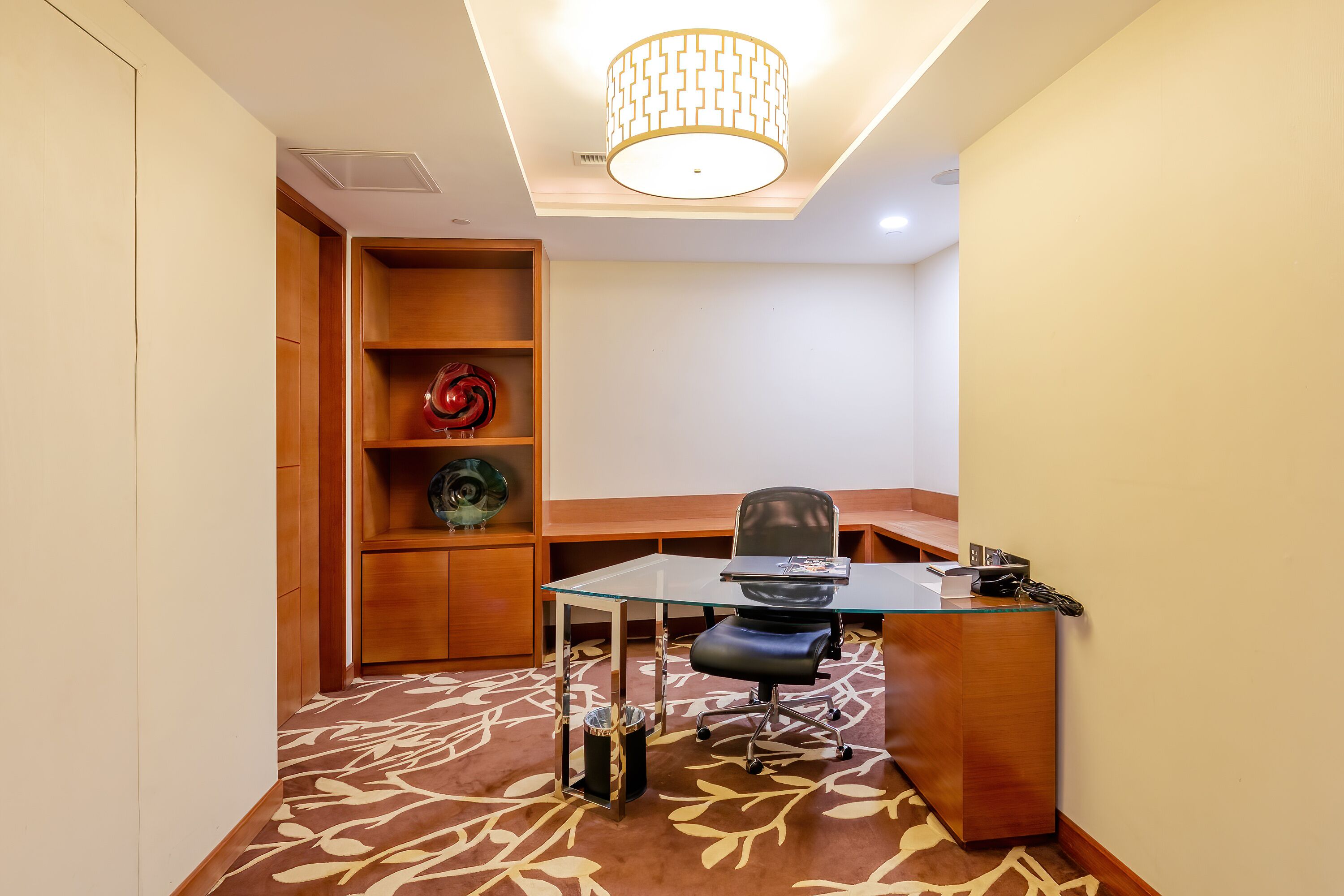 A modern office space with a glass desk, black office chair, and a wooden shelving unit with decorative items.